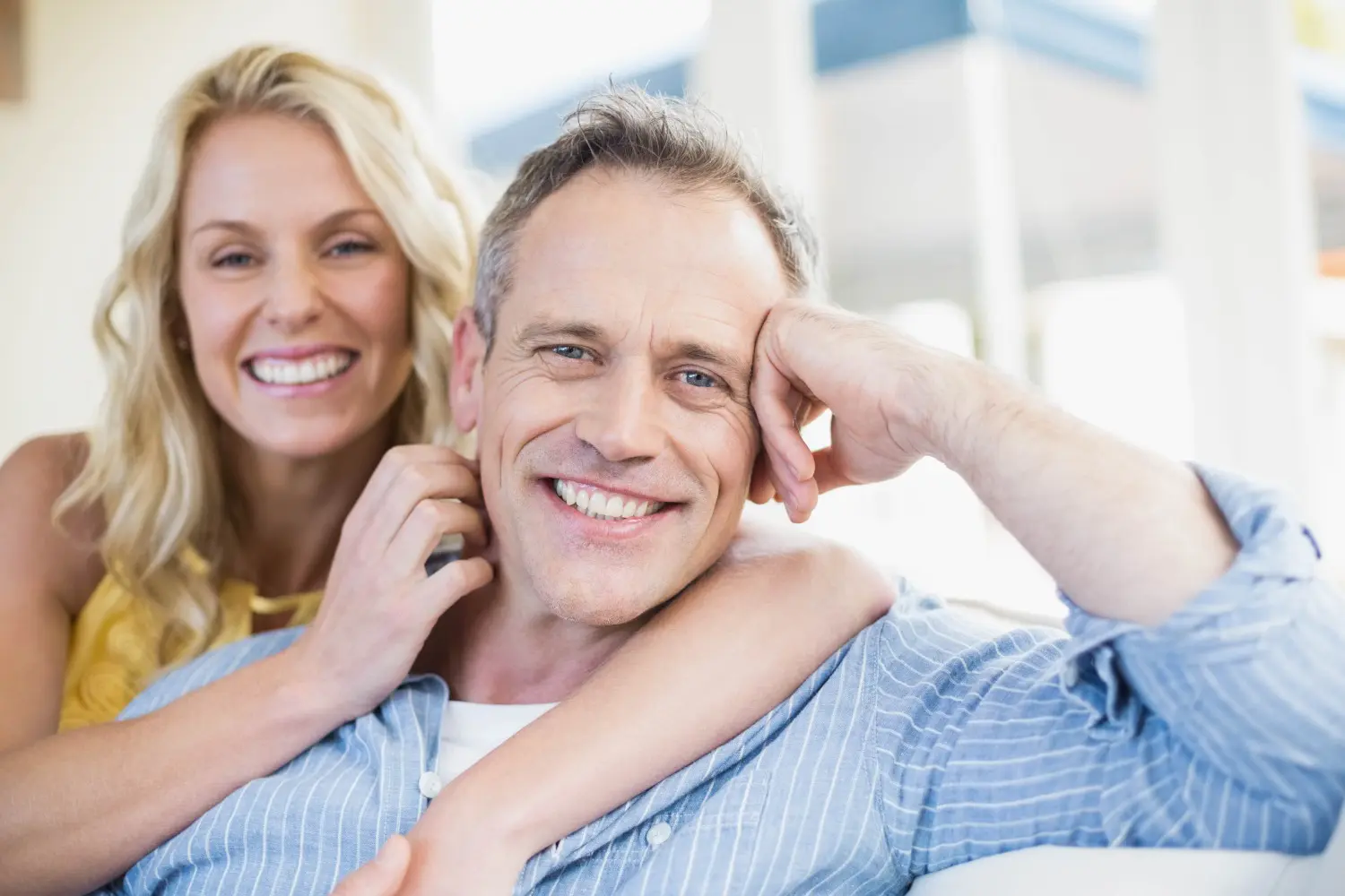 A smiling woman stands behind a man sitting on a sofa, her arm around his shoulders, as they review their Invisalign insurance options together in Suwanee or Tucker, GA