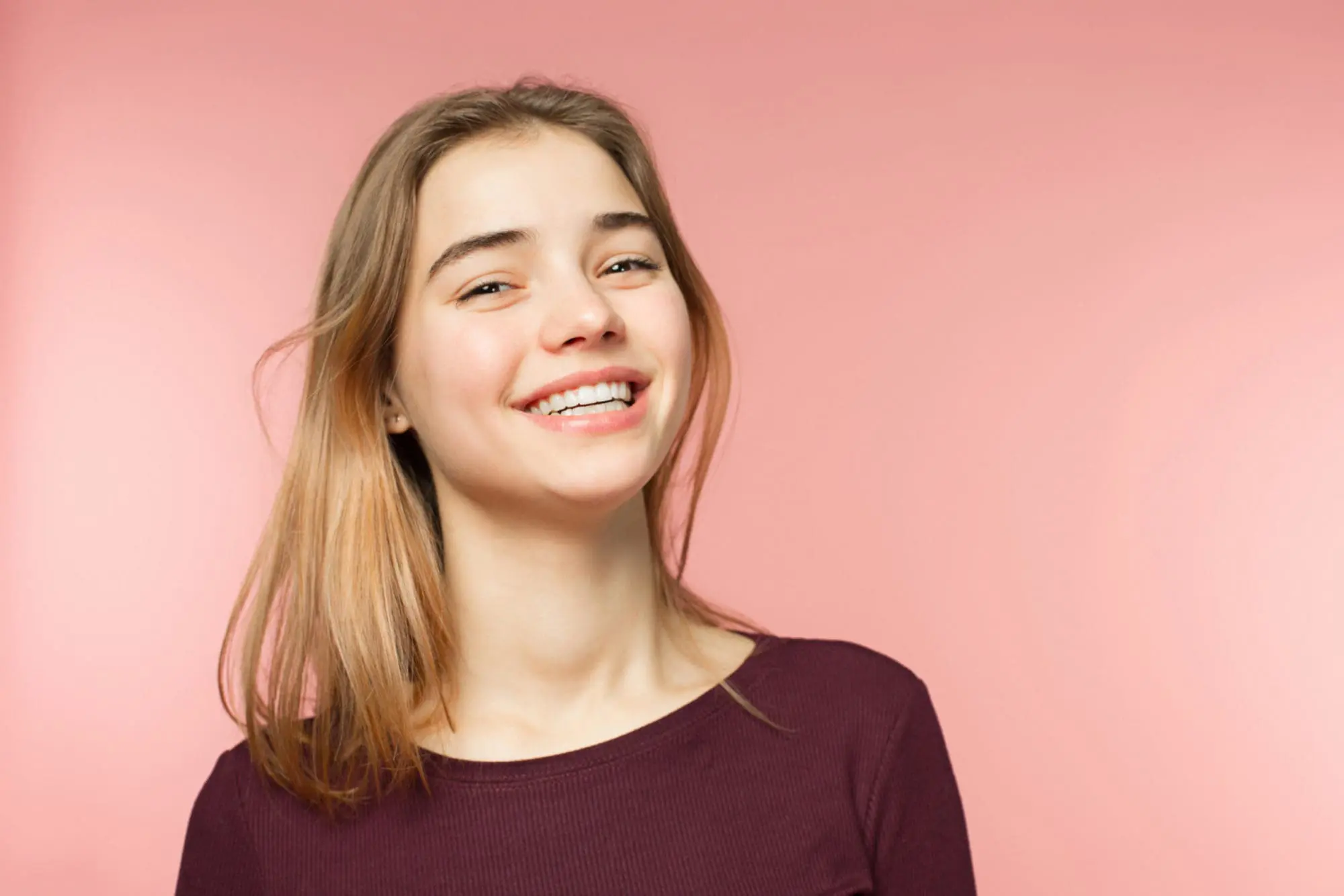 A person with shoulder-length blonde hair smiles confidently in front of a pink background, showcasing their beautifully aligned teeth thanks to NK Orthodontics, while wearing a dark long-sleeve shirt in Tucker, GA