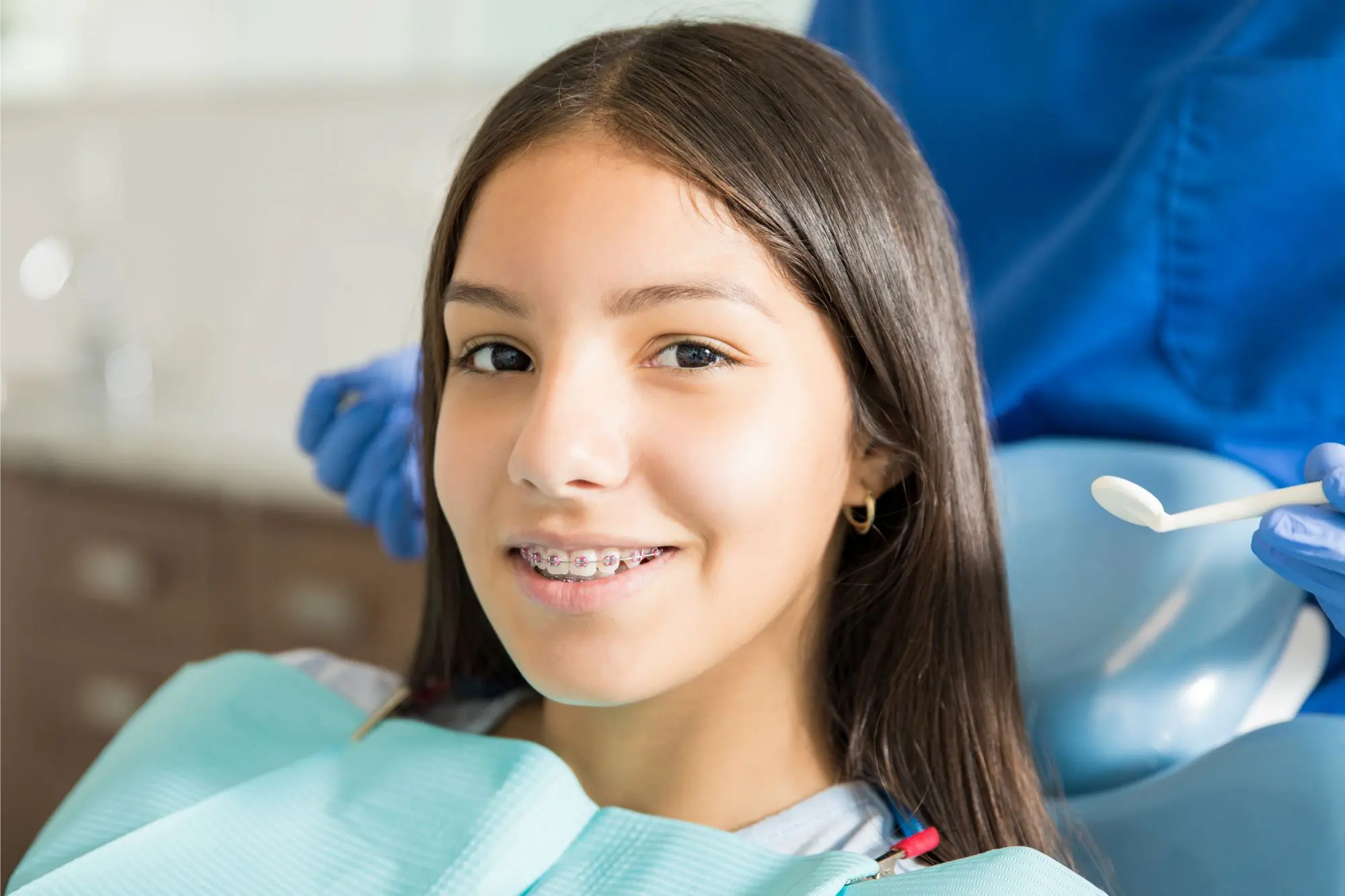A young person with braces sits in a dental chair at NK Orthodontics, wearing a dental bib. A dentist in blue gloves holds a dental tool nearby in Suwanee, GA.