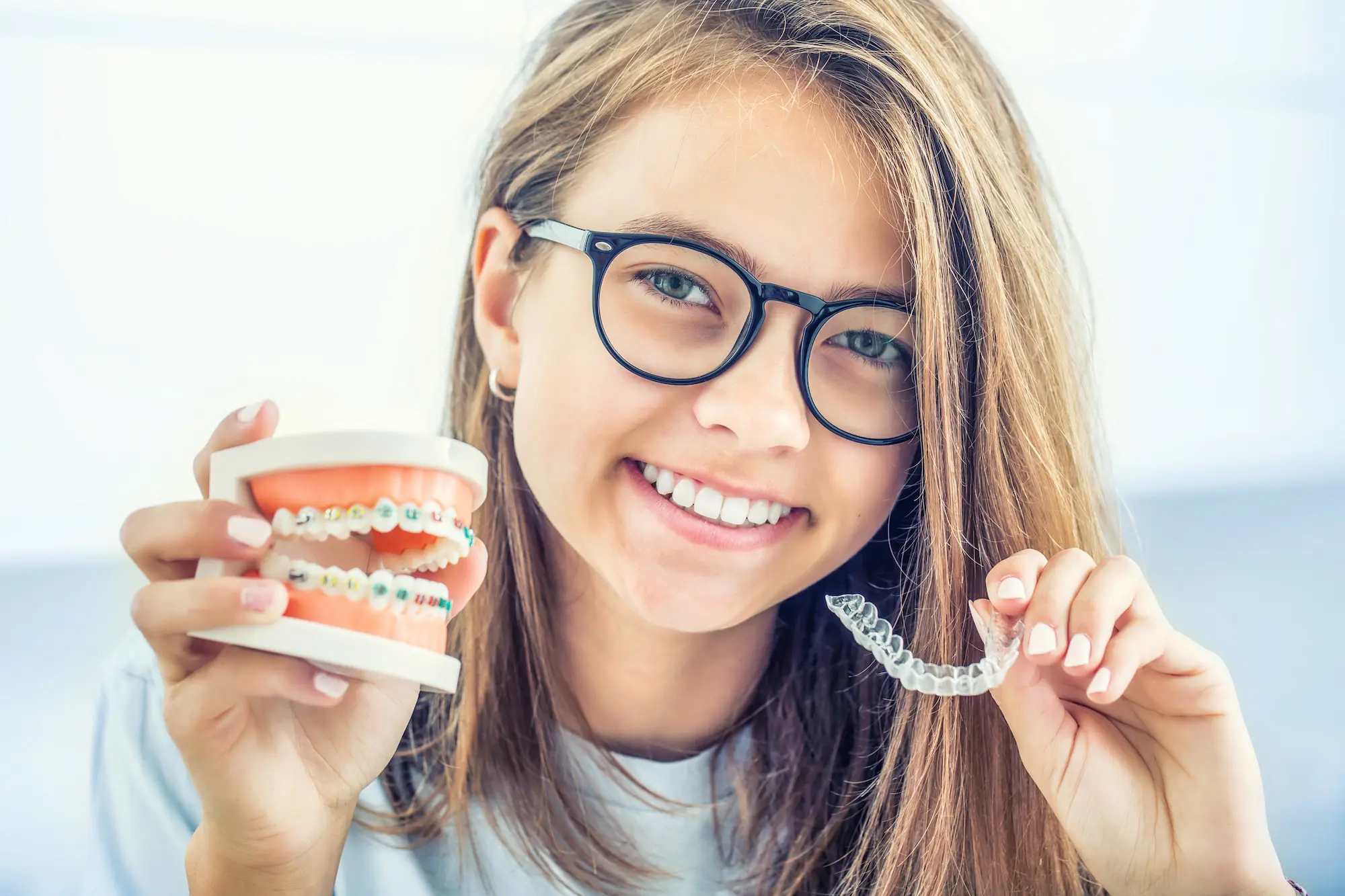 A person with glasses smiles while showcasing a dental model with braces in one hand and a clear aligner from NK Orthodontics in the other in Suwanee dan Tucker, GA