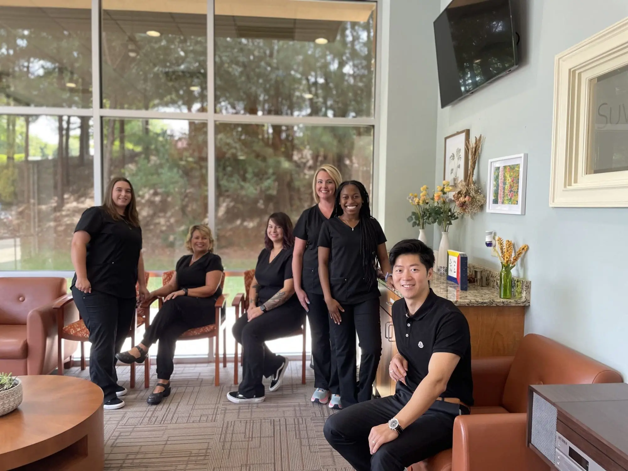Five people in black clothing are posing and sitting in a room with large windows, brown chairs, and a few decorative items on a cabinet at NK Orthodontics, where Dr. Nick Kim ensures every detail is perfectly aligned in Suwanee, GA