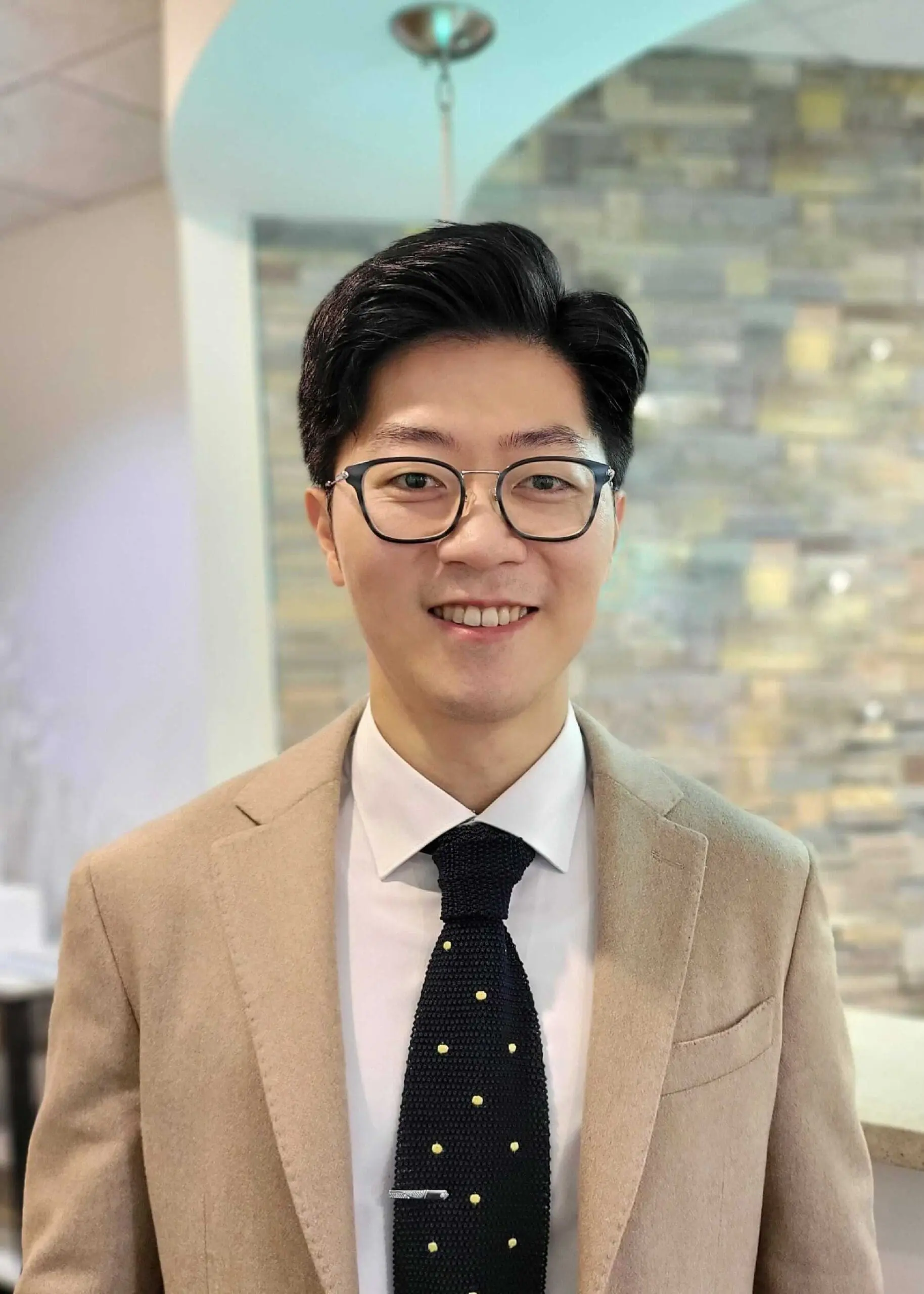 Dr. Nick Kim, sporting glasses, a beige suit jacket, and a dotted black tie, confidently poses in front of a stone wall—an emblematic image for NK Orthodontics in Suwanee, GA