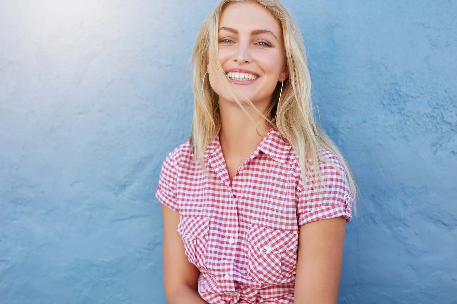 A woman in a red and white checkered shirt smiles in front of a blue wall, embodying the cheerful spirit of NK Orthodontics. Her blonde hair is partially covering her face, adding a touch of mystery to her joyful expression in Suwanee, GA.