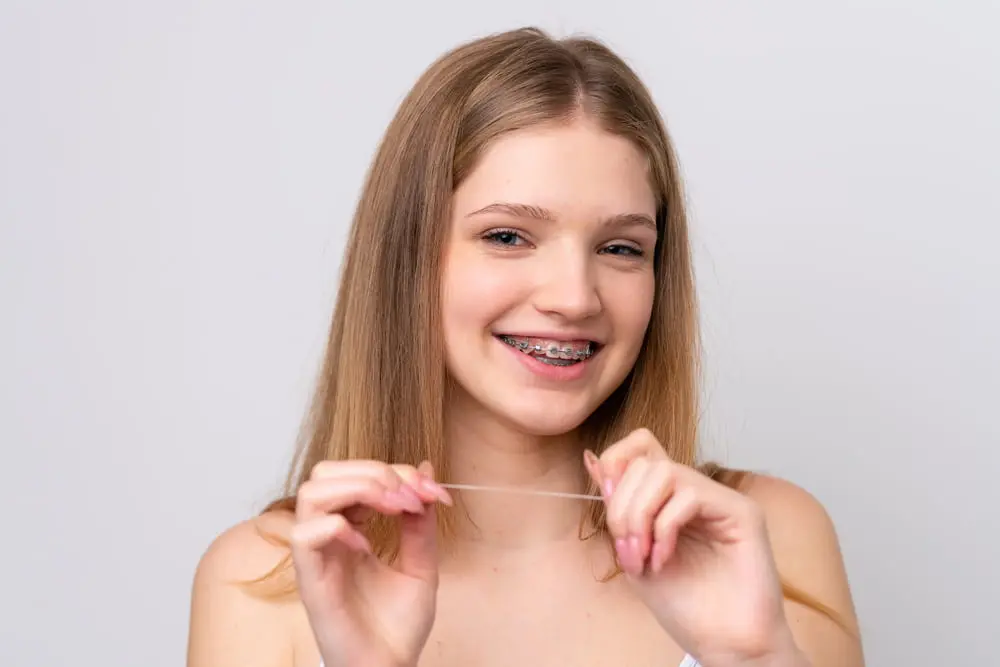 A young person with long hair and mtal braces beams as they hold up dental floss, proudly showcasing their commitment to oral health, thanks to NK Orthodontics, all set against a plain background in Suwanee or Tucker, GA