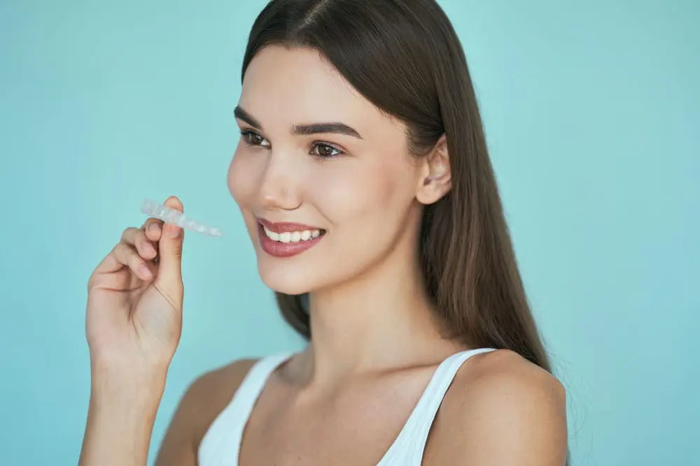 A woman with long brown hair smiles while holding a clear orthodontic aligner near her face against a light blue background, showcasing her experience with NK Orthodontics in Tucker, GA