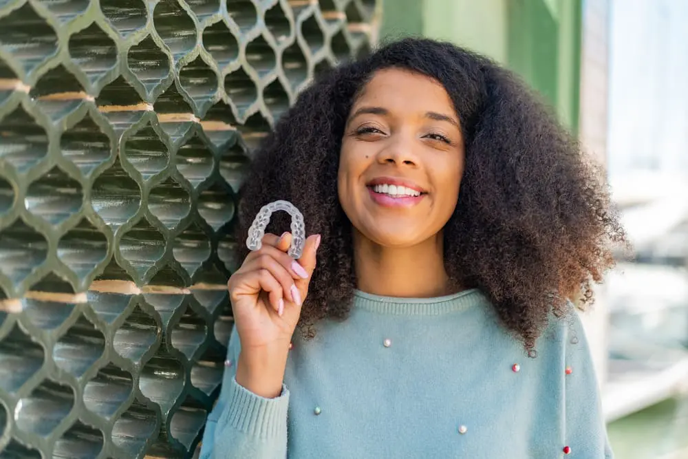 A woman beams brightly, holding a dental retainer, as she stands outdoors against a textured wall, showcasing her NK Orthodontics success in Suwanee, GA.