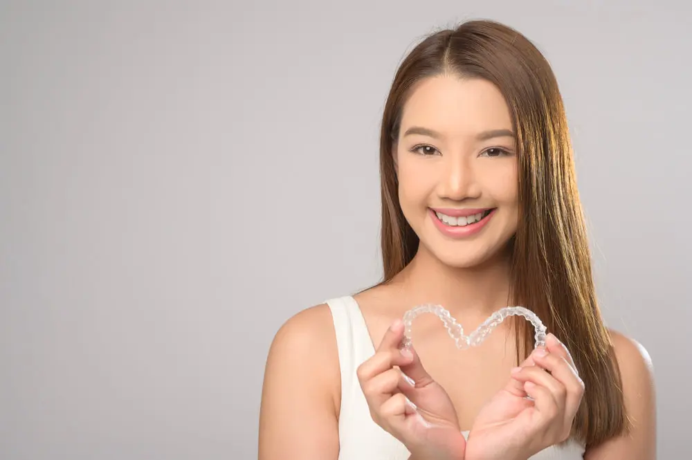 A woman with long hair beams as she holds an Invisalign aligner shaped like a heart against a plain background, showcasing the joy of adult orthodontics in Suwanee or Tucker, GA