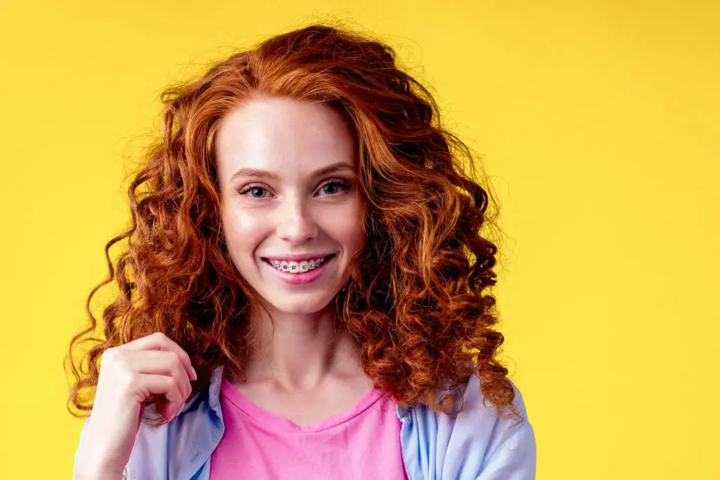 An adult with curly red hair and braces beams against a vibrant yellow background, donning a pink top and light blue jacket in Suwanee or Tucker, GA