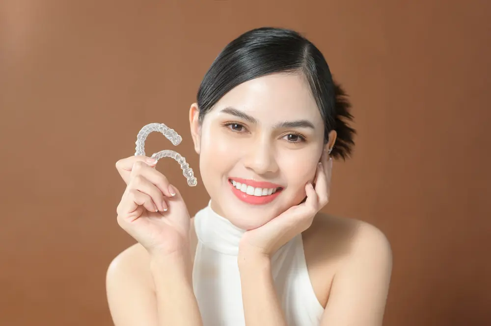 A woman, smiling confidently against a brown background, holds clear dental aligners. Wearing a white sleeveless top, she seems pleased with her choice despite the Invisalign cost in Suwanee or Tucker, GA