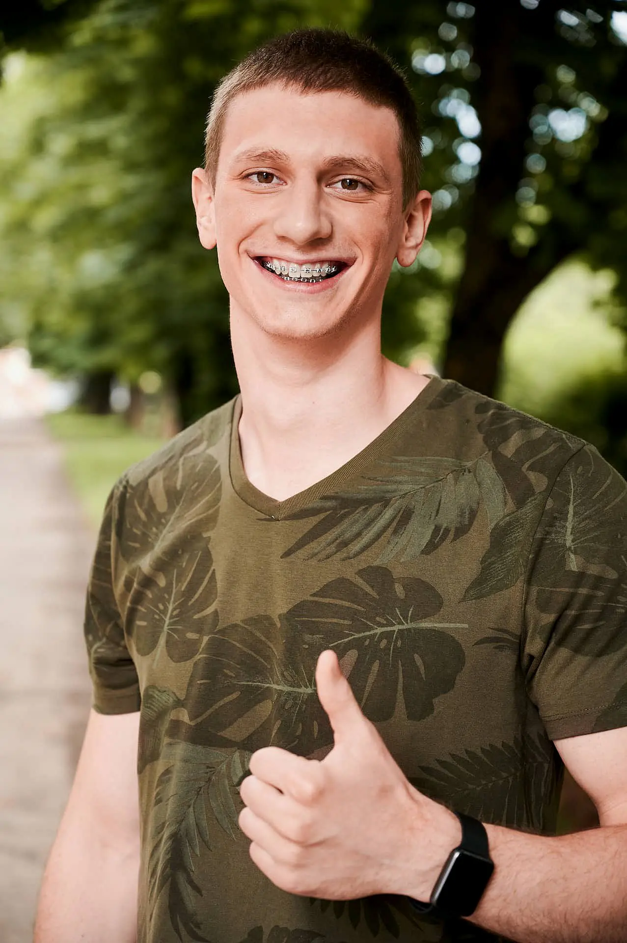A person with short hair and braces from adult orthodontics smiles, giving a thumbs up. They are wearing a green leafy patterned shirt and a watch, standing on a tree-lined path in Suwanee or Tucker, GA