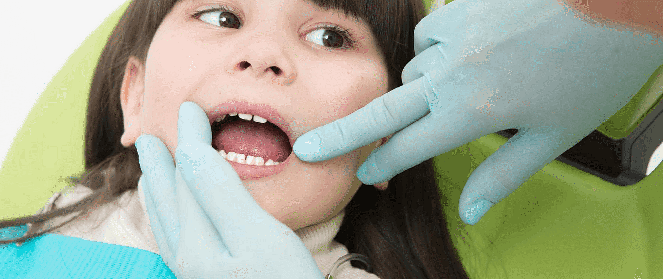 A child is sitting in a dental chair with their mouth open while a children's orthodontist wearing blue gloves carefully examines their teethin Suwanee or Tucker, GA