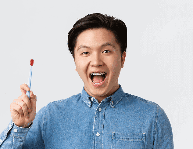 A person in a denim shirt, showcasing the beauty of adult orthodontics, holds a blue toothbrush with red bristles while smiling broadly against a plain background in Suwanee or Tucker, GA