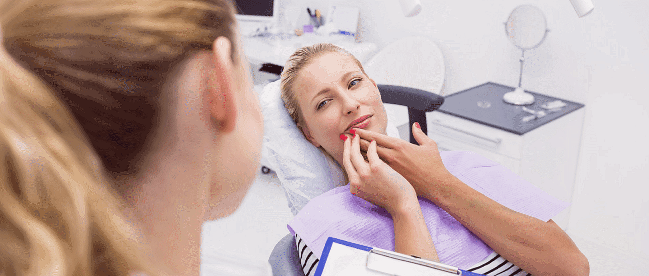 A woman lies in a dental chair, touching her lip and looking at a female dentist who is holding a clipboard, discussing options in adult orthodontics in Suwanee or Tucker, GA