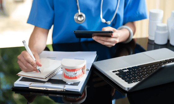 A person in blue scrubs writes in a notebook while holding a smartphone, likely reviewing orthodontic insurance details. Nearby are a dental model, a laptop, and medical bottles on the desk in Suwanee or Tucker, GA