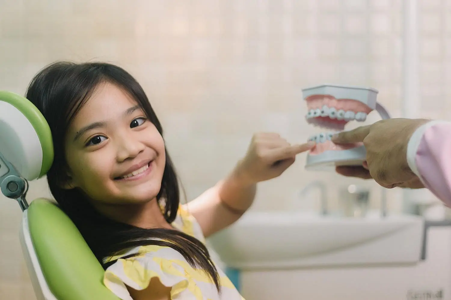 A young girl in a dental chair smiles and points at a dental model held by a person in a white coat, showcasing the expertise of children's orthodontists in a friendly dentist's office setting in Suwanee or Tucker, GA