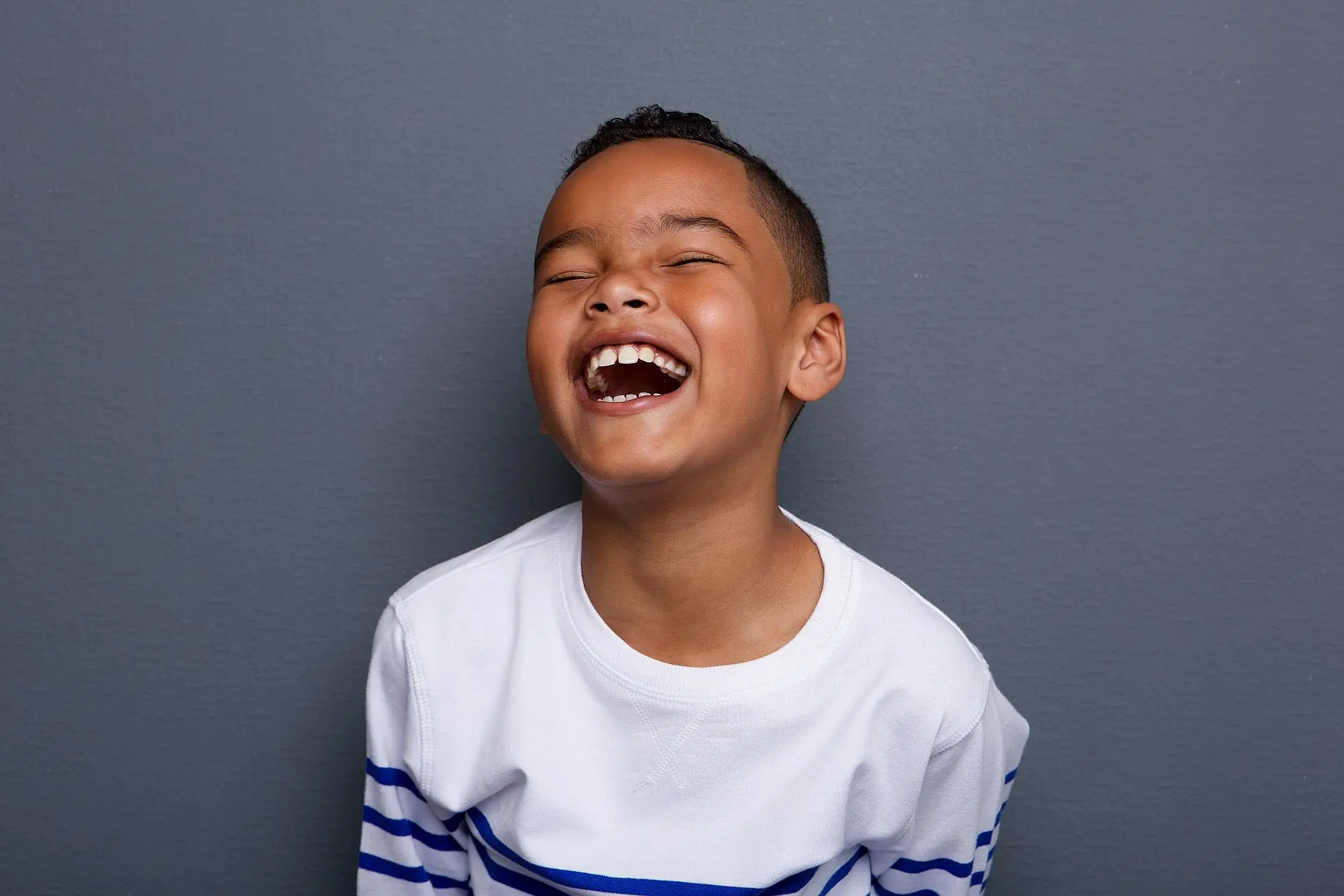 A child in a white shirt with blue stripes is laughing joyfully against a gray wall, showcasing the confidence that can come from regular visits to a children’s orthodontist in Suwanee or Tucker, GA