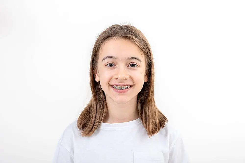 A young person with long brown hair and braces beams at the camera, showcasing the expert work of children's orthodontists. Wearing a white t-shirt, they stand confidently against a plain white background in Suwanee or Tucker, GA