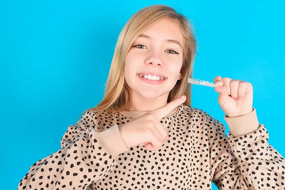 A girl, smiling in a spotted shirt, happily points at clear dental aligners against a blue background, embodying the expertise of a children's orthodontist in Suwanee or Tucker, GA