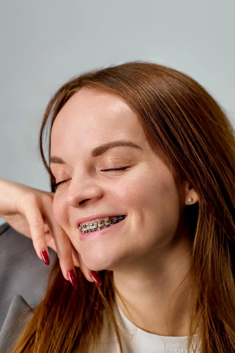 A woman with long brown hair and braces, showcasing the benefits of adult orthodontics, smiles with closed eyes, resting her cheek on her hand in Suwanee or Tucker, GA