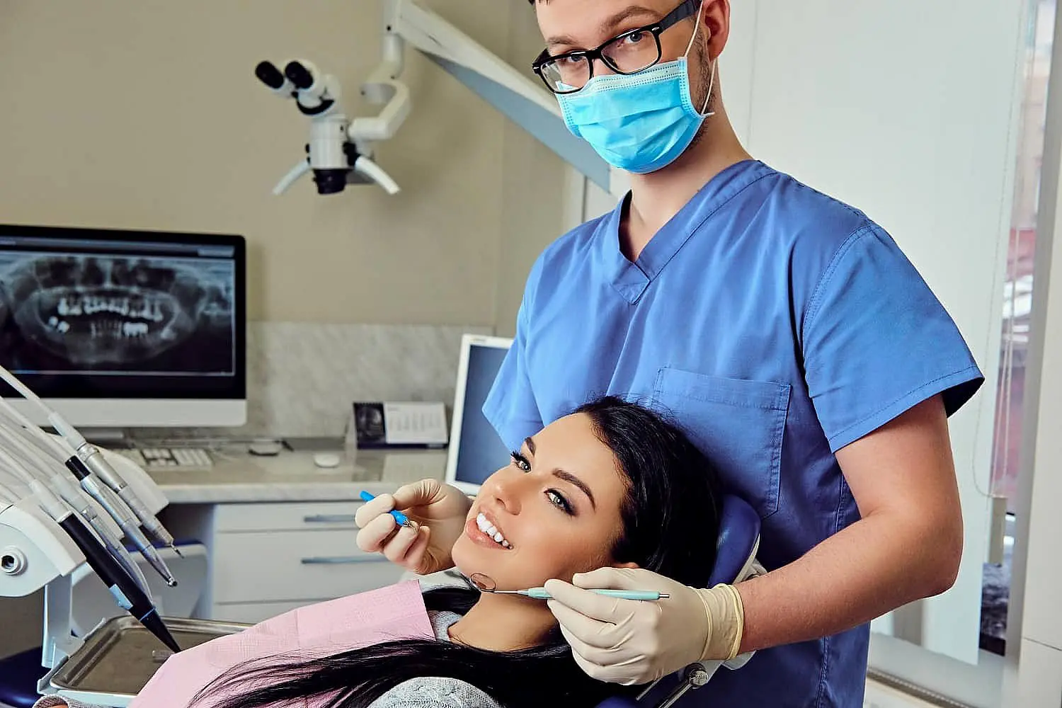 A dentist in blue scrubs and mask holds dental tools, including a palatal expander, while standing next to a patient reclining in a chair, with dental equipment and an X-ray on a screen in the background in Suwanee or Tucker, GA