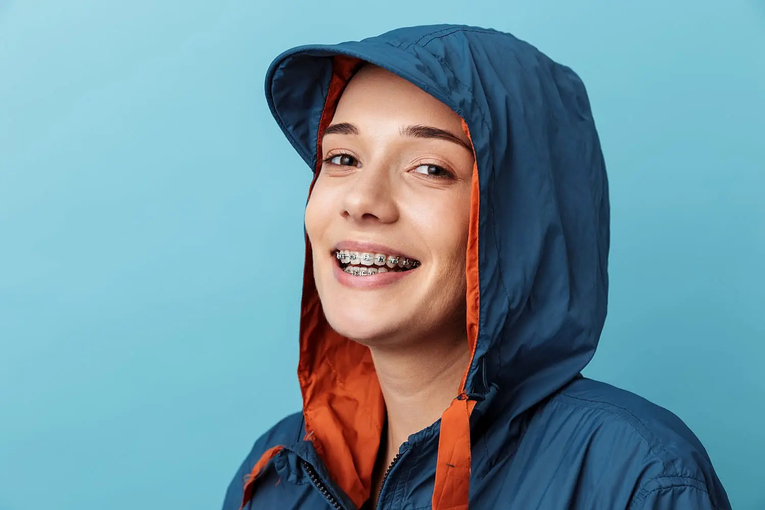 A person, sporting an overbite, smiles brightly with braces on their teeth while wearing a blue and orange hooded jacket against a vibrant blue background in Suwanee or Tucker, GA