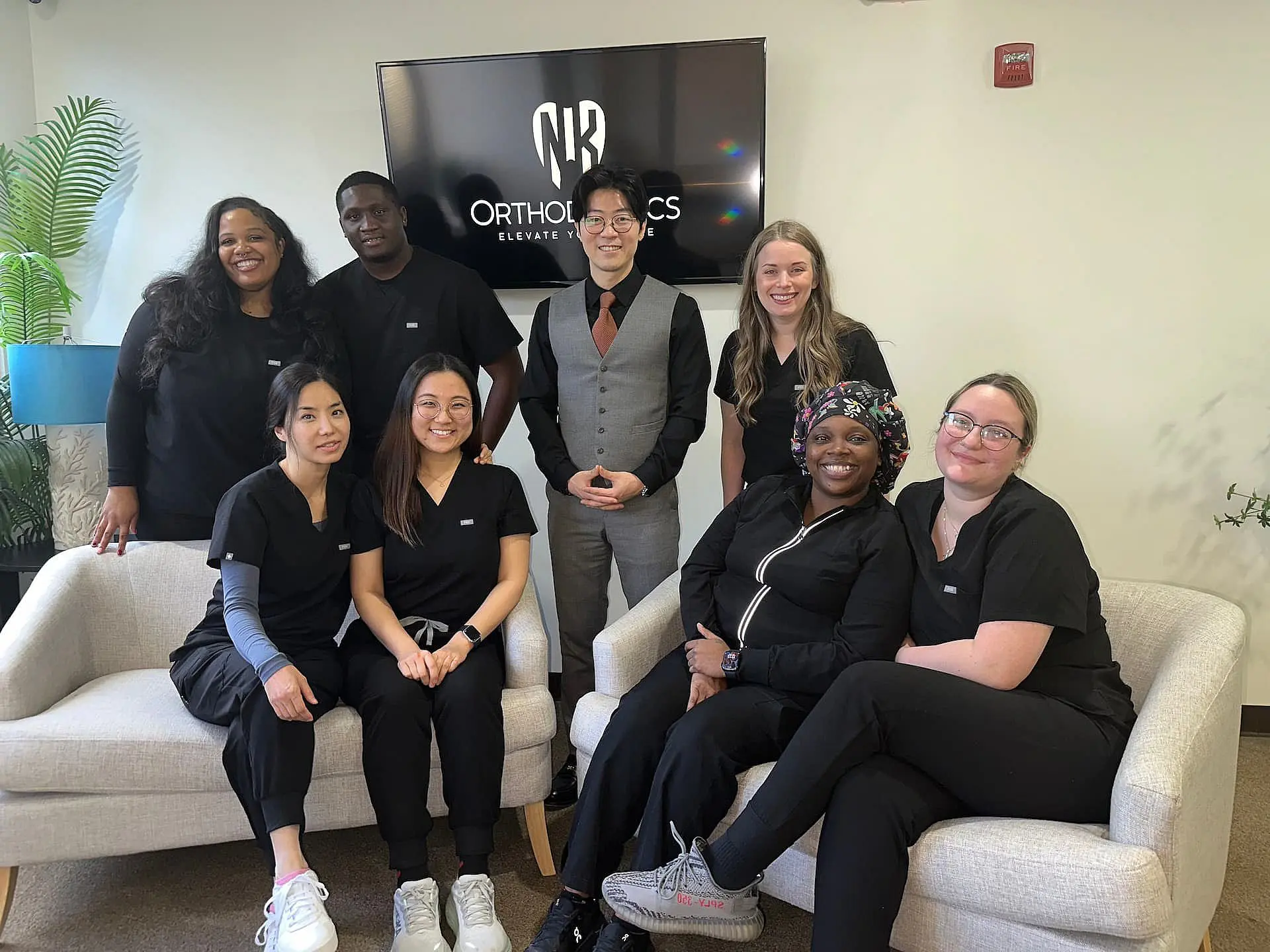 A group of eight people, mostly clad in black, pose in an office setting with a sign that reads "Orthodontics" in the background. Two are seated on sofas while the others stand behind. They appear ready to discuss orthodontic insurance options with clients in Suwanee or Tucker, GA