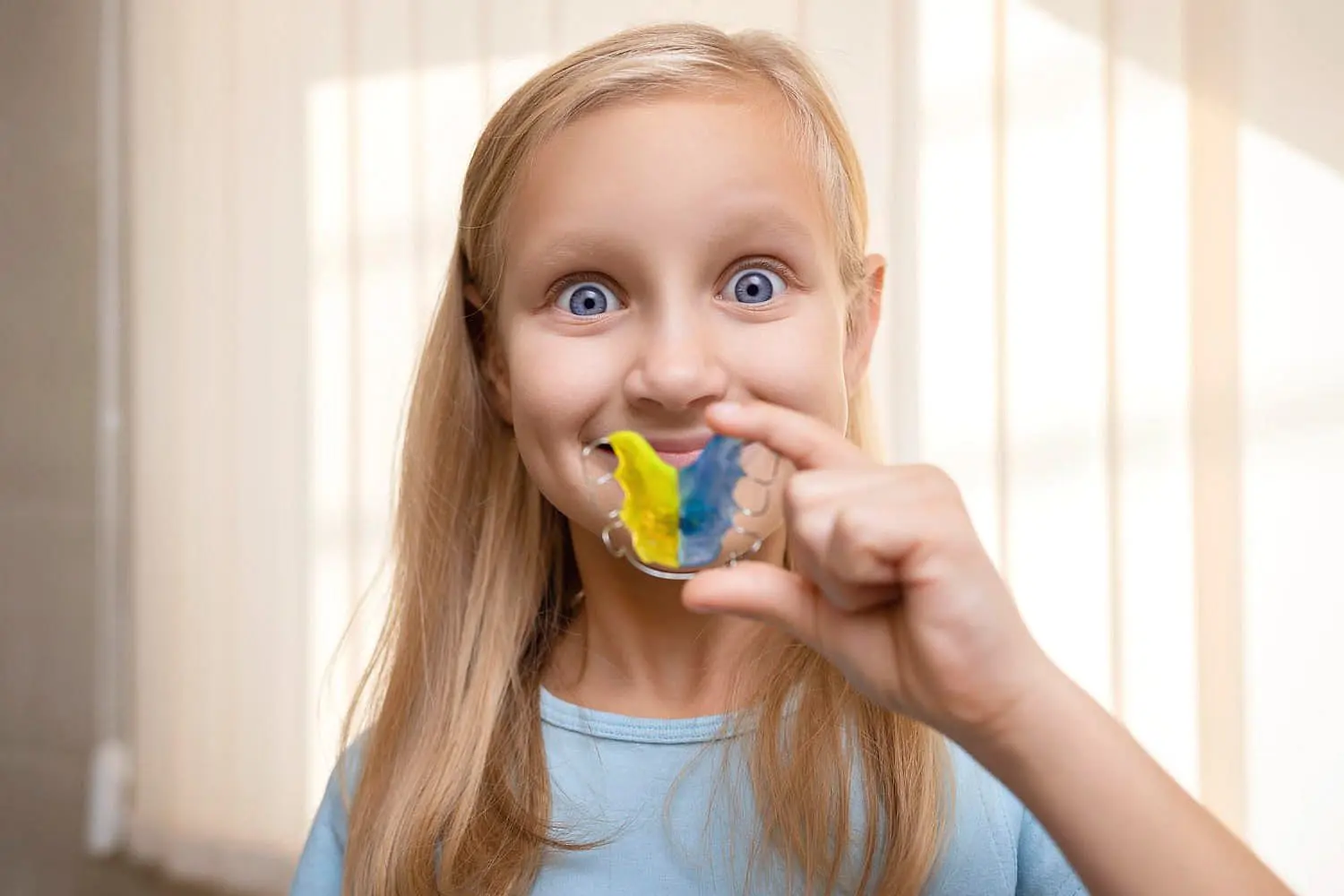 A child happily holds an orthodontic appliance, a vibrant mix of blue and yellow, in front of their face while flashing a big smile in Suwanee and Tucker, GA