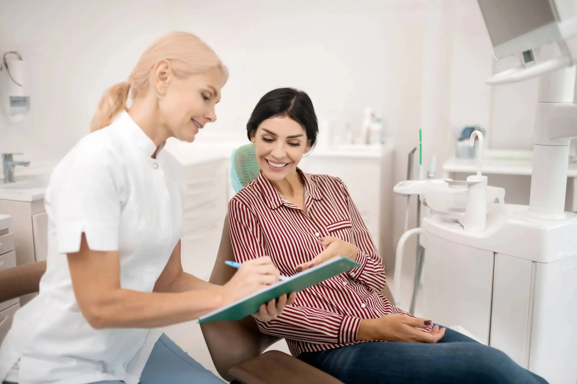 A dentist shows a clipboard to a smiling woman sitting in a dental chair, discussing her customized treatment plan. In the cozy dental office, equipped with state-of-the-art tools, they explore the benefits of affordable Invisalign for achieving that perfect smilein Suwanee or Tucker, GA