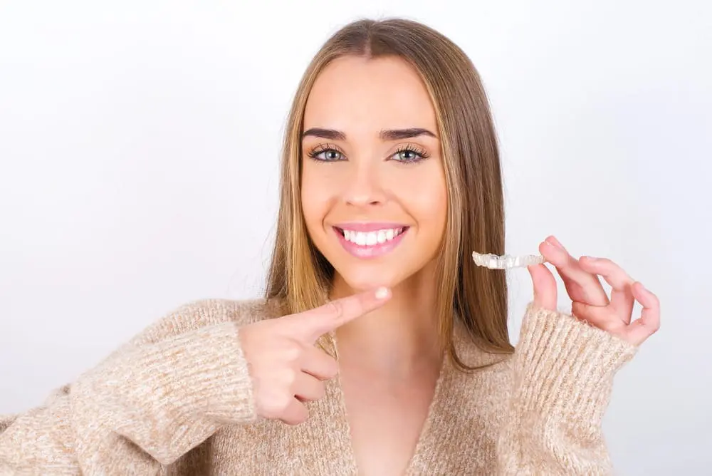 Smiling woman in a beige sweater holds an Invisalign® Clear Aligner, proudly pointing to her teeth against a plain background, showcasing her journey with NK Orthodontics in Suwanee or Tucker, GA