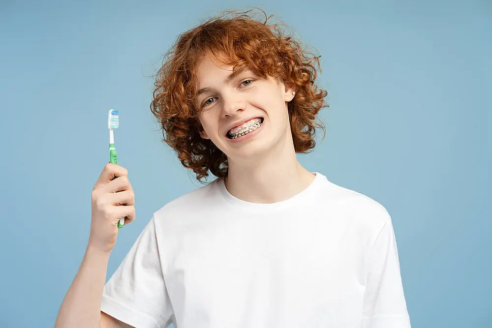 A teen with red curly hair smiles brightly, showcasing his metal braces while holding a toothbrush. Dressed in a white t-shirt against a light blue background, he radiates confidence and charm in Tucker, GA