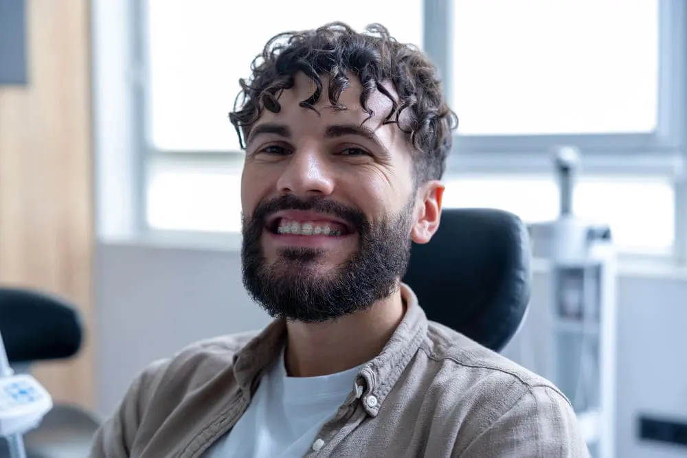A smiling man with curly hair and a beard, sporting ceramic braces, is seated in a brightly lit room in Suwanee, GA