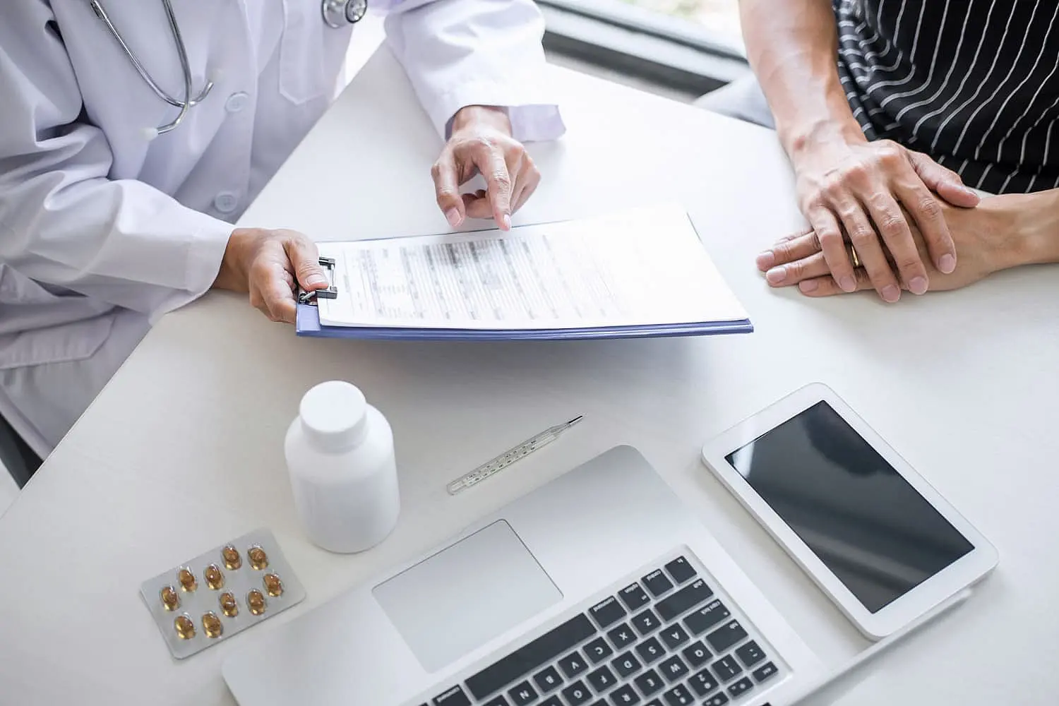 The doctor, specializing in orthodontic insurance, reviews a medical form with the patient at a desk. The setup includes a laptop, tablet, medication bottle, pills, and a thermometer, ensuring comprehensive care in Suwanee or Tucker, GA