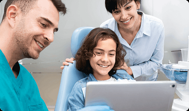 A child sits in a dental chair, smiling at a tablet held by the children’s orthodontist. An adult stands nearby, also smiling in Suwanee or Tucker, GA