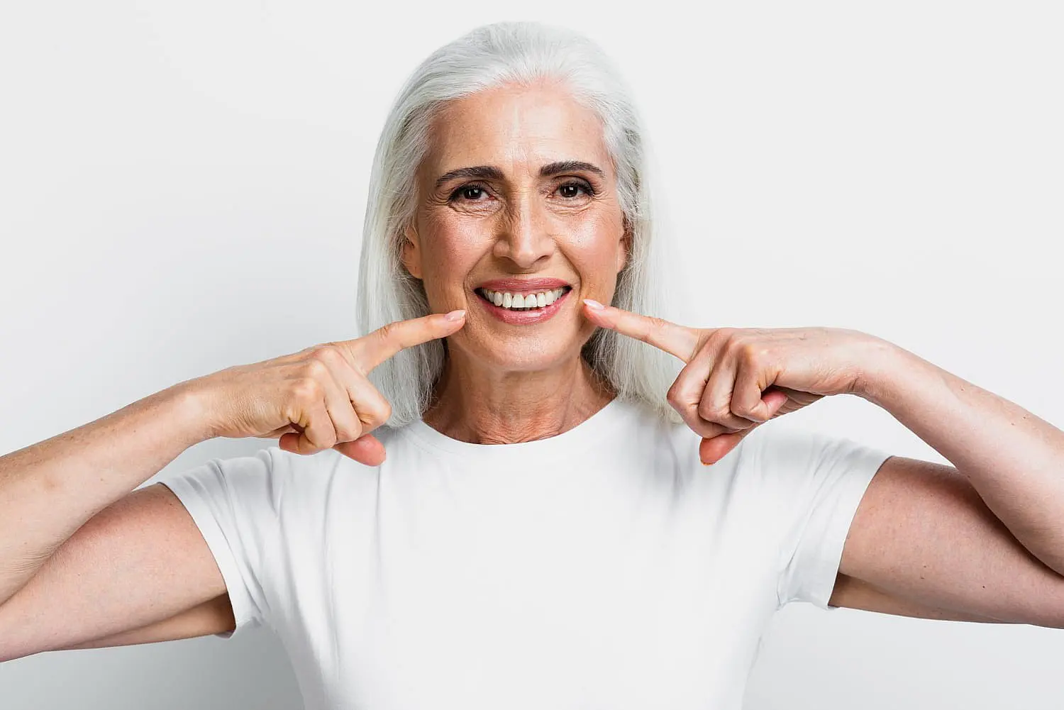 Smiling woman with long gray hair and a white shirt points at her cheeks, highlighting the confidence that comes with orthodontic insurance against a plain background in Suwanee or Tucker, GA