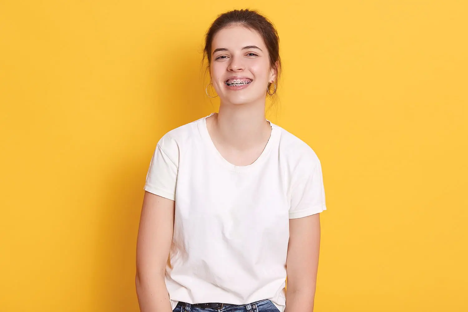 A person with an overbite, wearing braces and a white t-shirt, smiles warmly against a yellow background, hoop earrings glinting in the light in Suwanee or Tucker, GA