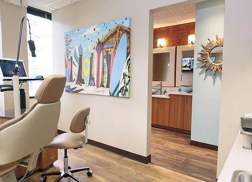 Children’s Orthodontists' office with a surfboard-themed painting on the wall, beige dental chair, and a view into an adjacent room with wooden cabinets and a sunburst mirror in Suwanee or Tucker, GA