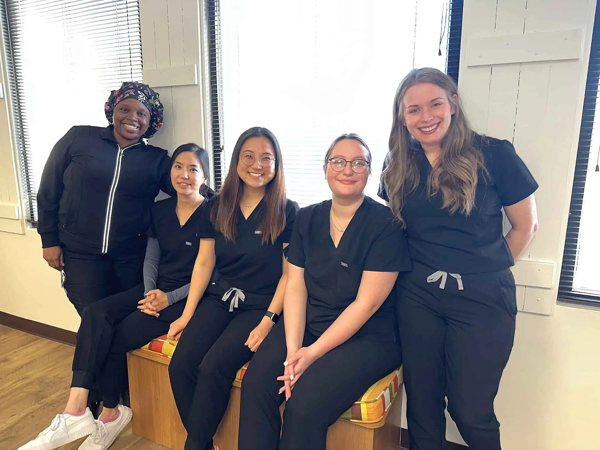 Five people in matching black scrubs, possibly dental professionals who can answer your Invisalign be covered by insurance questions, sit and stand in a brightly lit room with windows, smiling at the camera in Suwanee or Tucker, GA