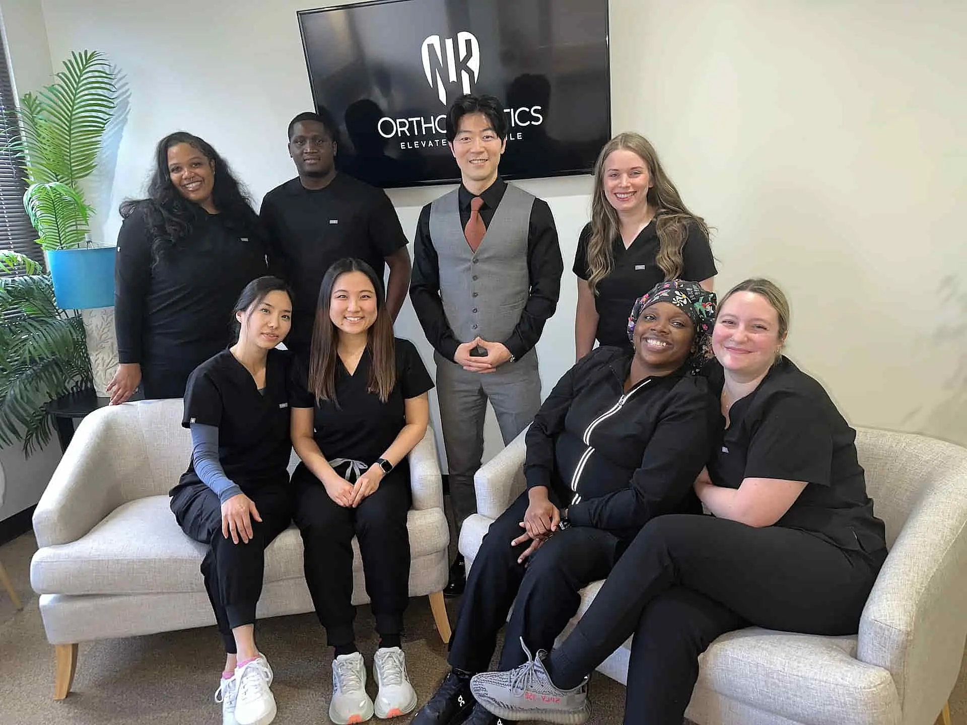 A group of eight people in black attire poses in an office with "Orthodontics" and "Adult Braces" displayed on a TV screen. Two are seated on couches, while the others stand around them, showcasing their professional demeanor in Suwanee or Tucker, GA