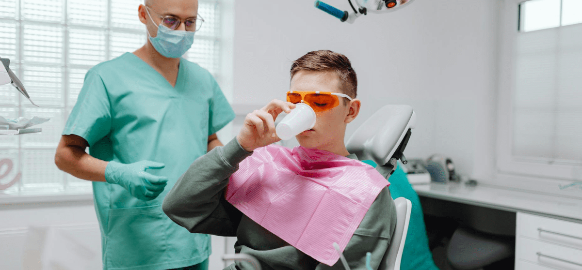 A boy sits in a dental chair at NK Orthodontics, wearing protective glasses and a bib, sipping from a cup. Beside him stands the dentist in green scrubs and gloves, ensuring his comfort in Suwanee or Tucker, GA