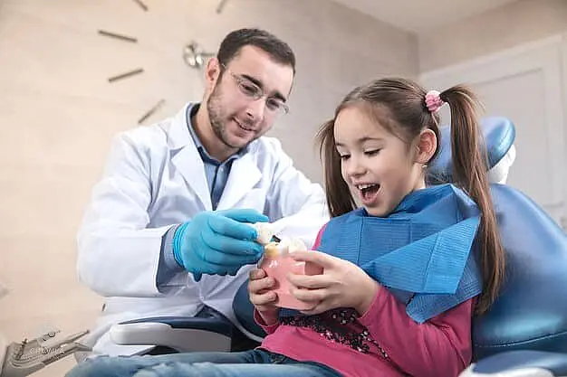 A children's orthodontist shows a dental model to a smiling young girl sitting in a dental chair, introducing her to the importance of retainers for a lifelong healthy smile in Suwanee or Tucker, GA