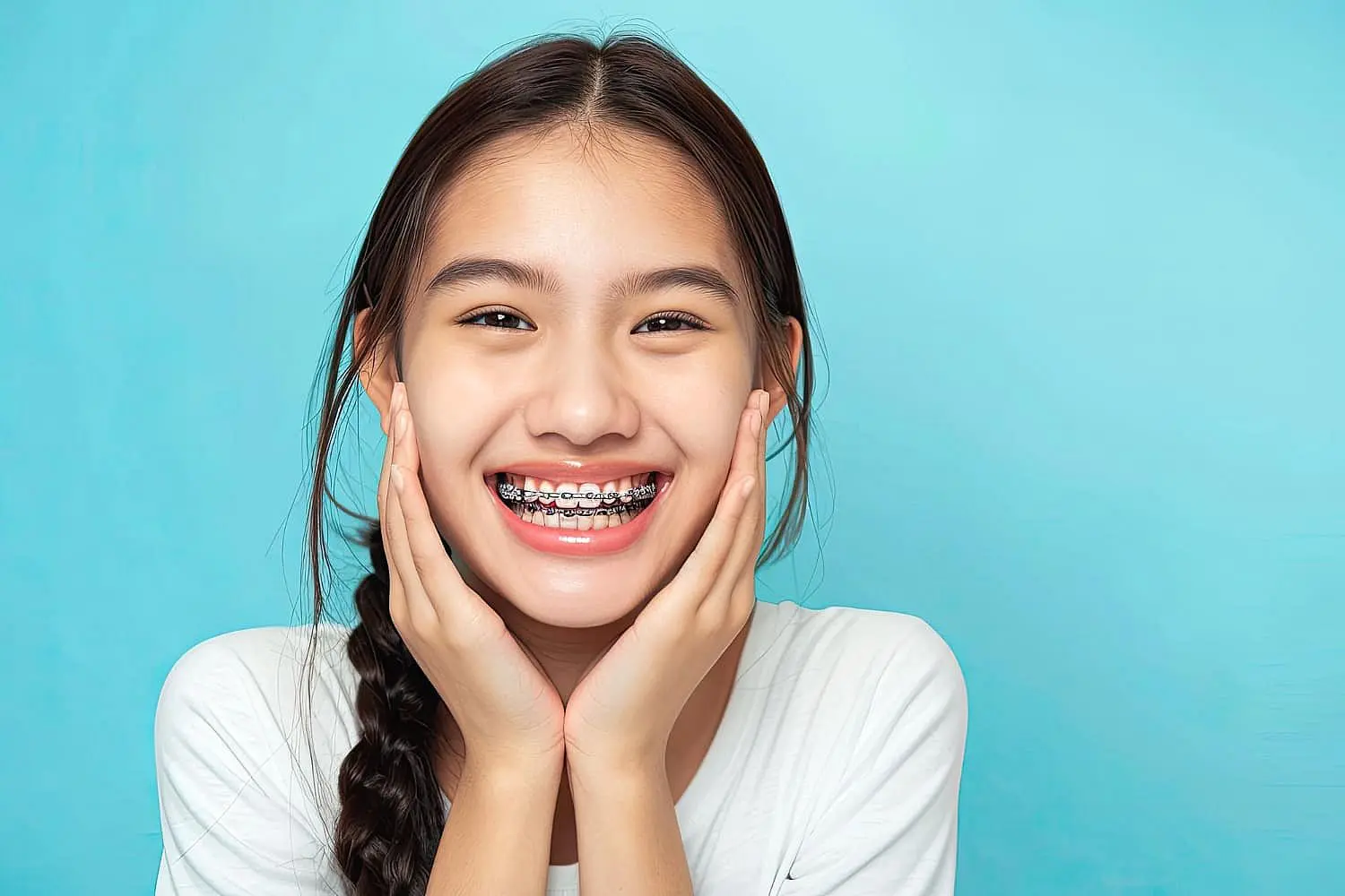 A smiling person with long hair and traditional braces, wearing a white shirt, holds their face with both hands against a blue background, showcasing the progress of their journey with a children's orthodontist in Suwanee or Tucker, GA