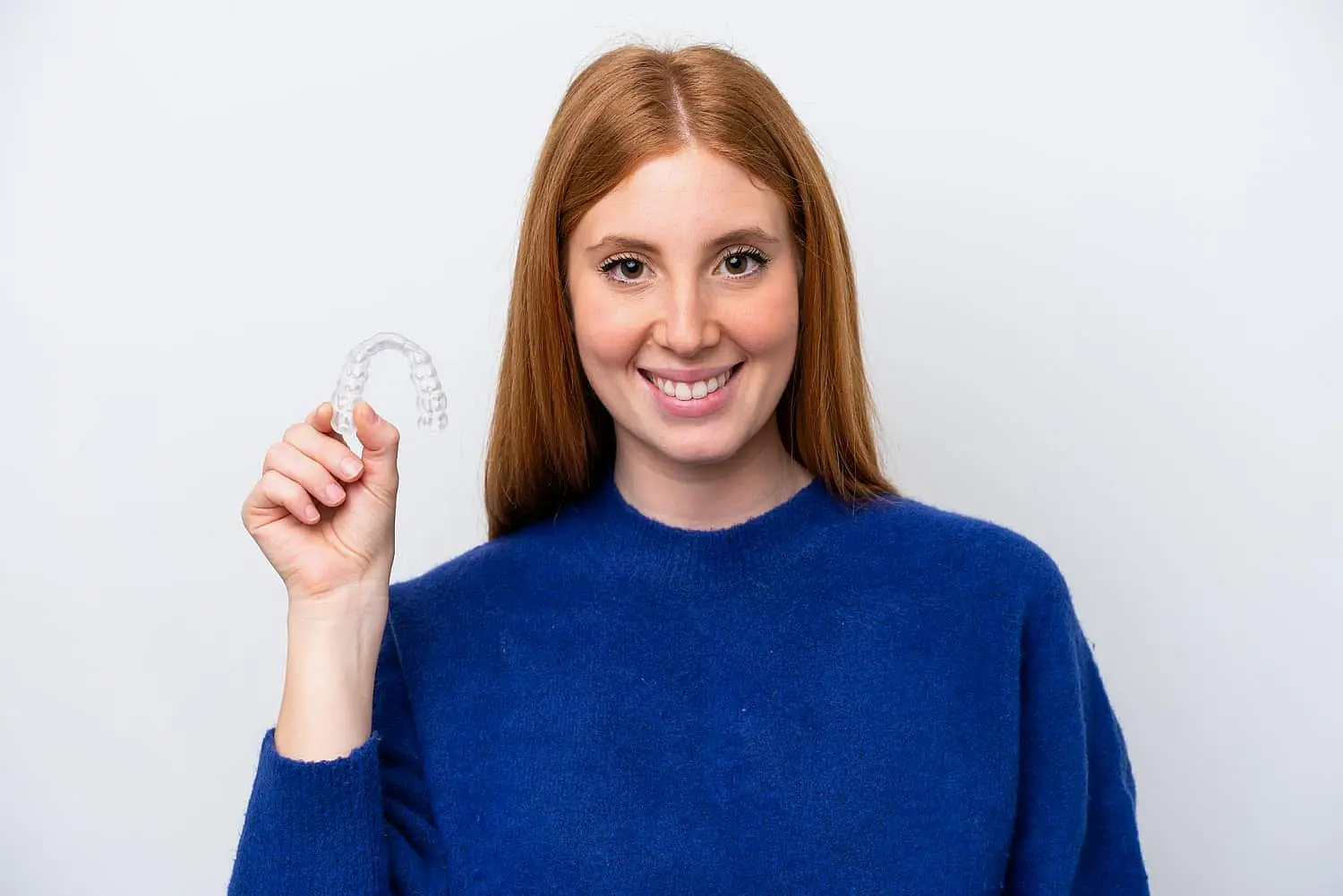 A woman with long red hair, wearing a blue sweater, smiles as she holds up an affordable Invisalign aligner against a plain background in Suwanee or Tucker, GA