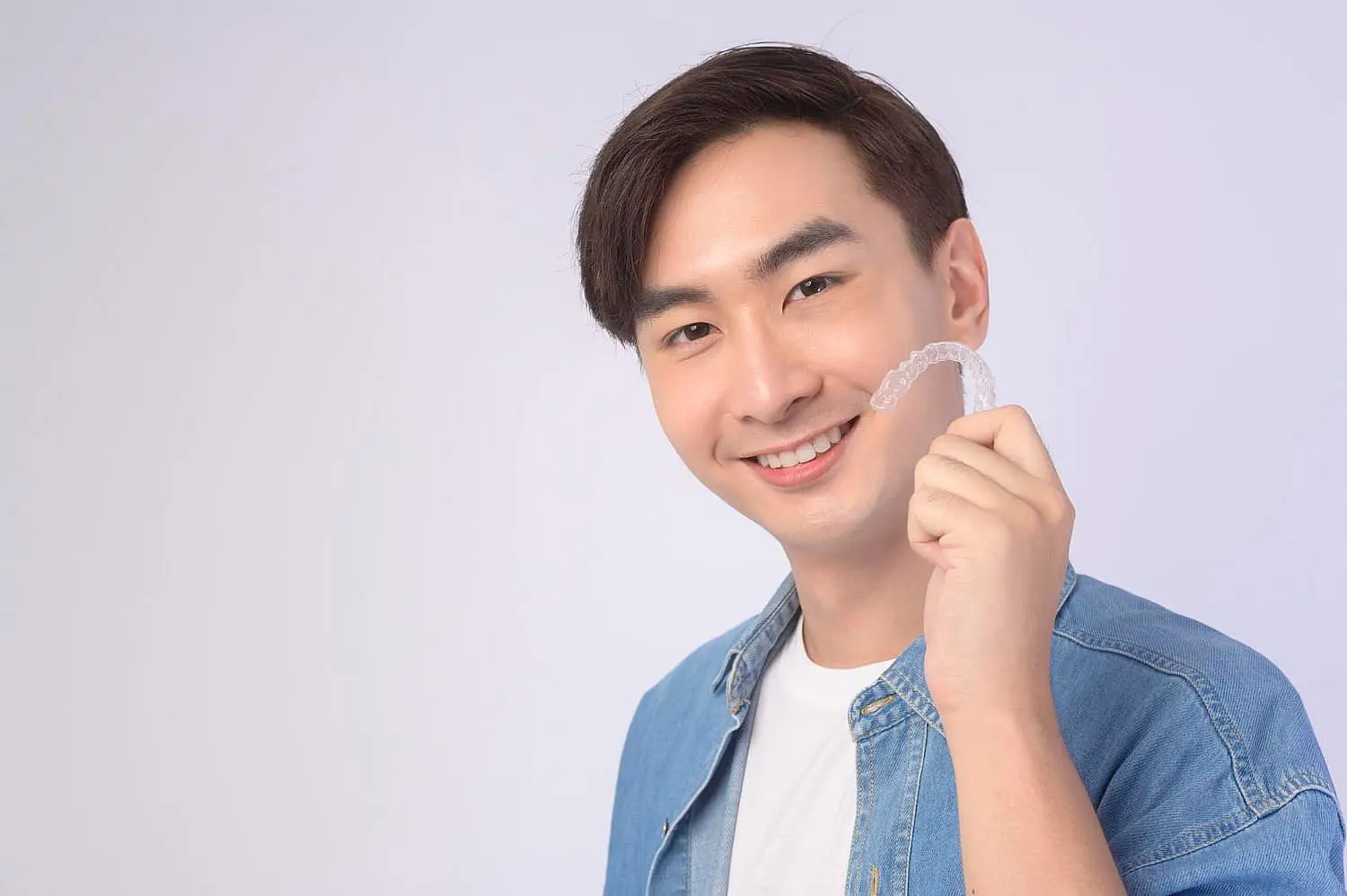 A person in a denim shirt smiles while holding a clear dental aligner against a light gray background, showcasing their bright smile as they appreciate the affordable Invisalign cost in Suwanee or Tucker, GA
