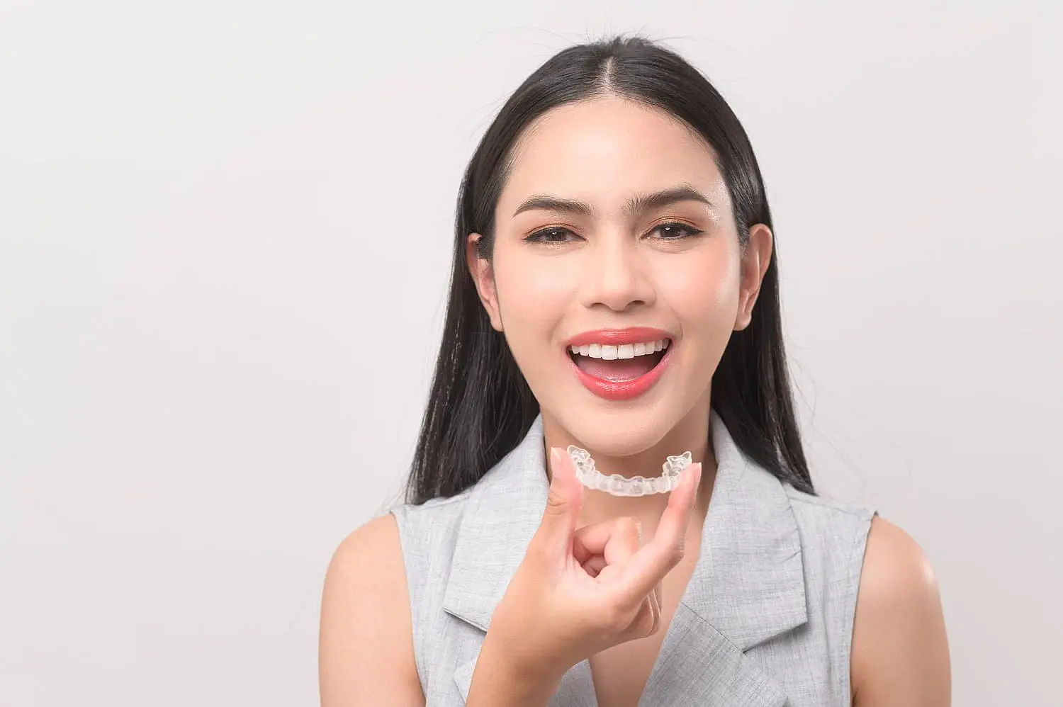 A person with long dark hair smiles while holding an Invisalign clear aligner near their mouth. They are wearing a sleeveless gray top in Tucker, GA