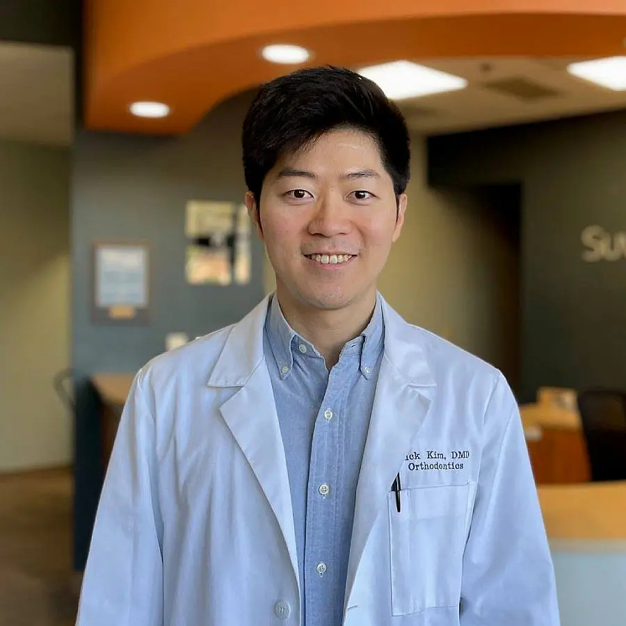 dr-nick-headshot-update A smiling man in a dental office, Dr. Nick Kim, wearing a white coat and orange tie, stands confidently in front of a reception desk in Suwanee or Tucker, GA
