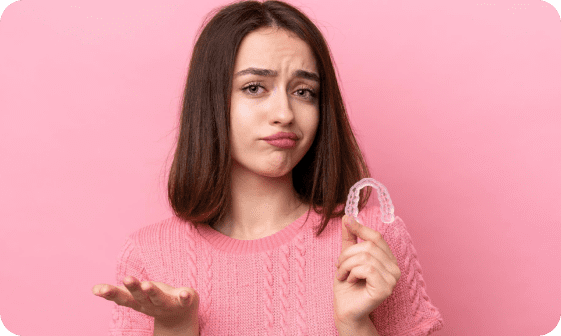 A woman in a pink sweater holds a dental retainer with a questioning expression against a pink background, pondering if her orthodontic insurance will cover the cost in Suwanee or Tucker, GA