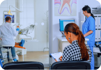 A woman in a checked shirt fills out an orthodontic insurance form in a dental office waiting area. In the background, a dentist attends to a patient in Suwanee or Tucker, GA
