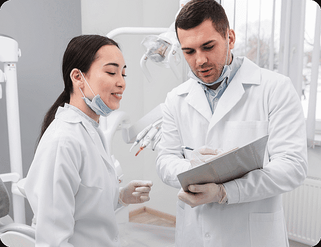 Two dental professionals in white coats discuss notes on a clipboard in a dental office, focusing on adult orthodontics while wearing masks and gloves. Dental equipment is visible in the background in Suwanee or Tucker, GA
