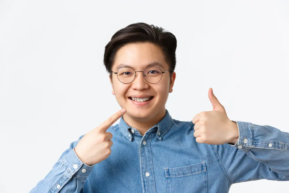 An adult wearing glasses and braces gives a thumbs-up and points at their mouth, smiling proudly. They are dressed in a denim shirt against a plain white background in Suwanee or Tucker, GA