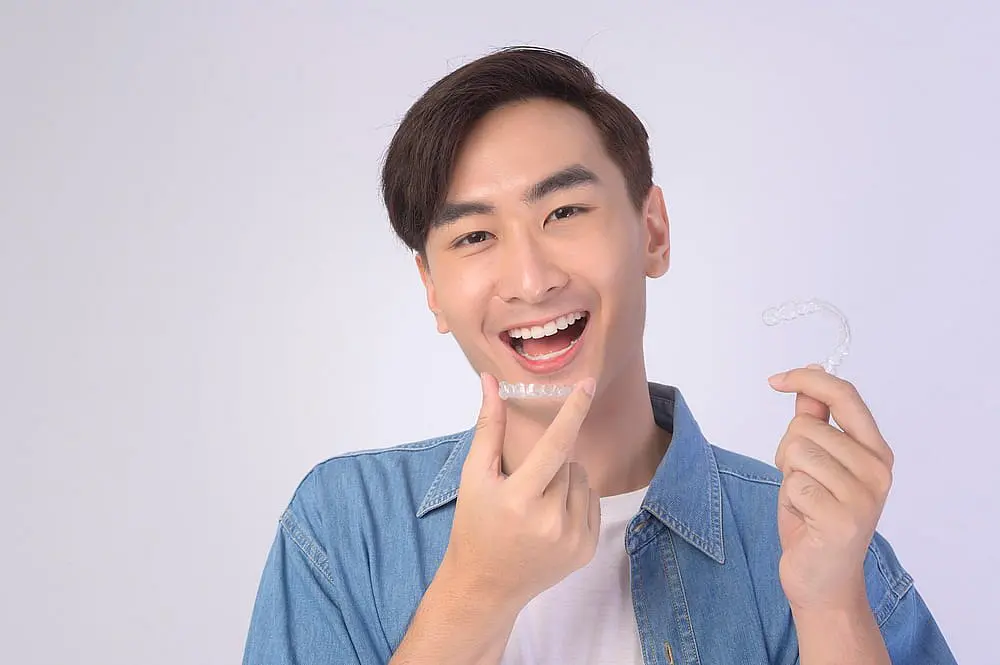 Person in a denim shirt smiles while holding a clear Invisalign aligner, about to place it in their mouth in Tucker, GA